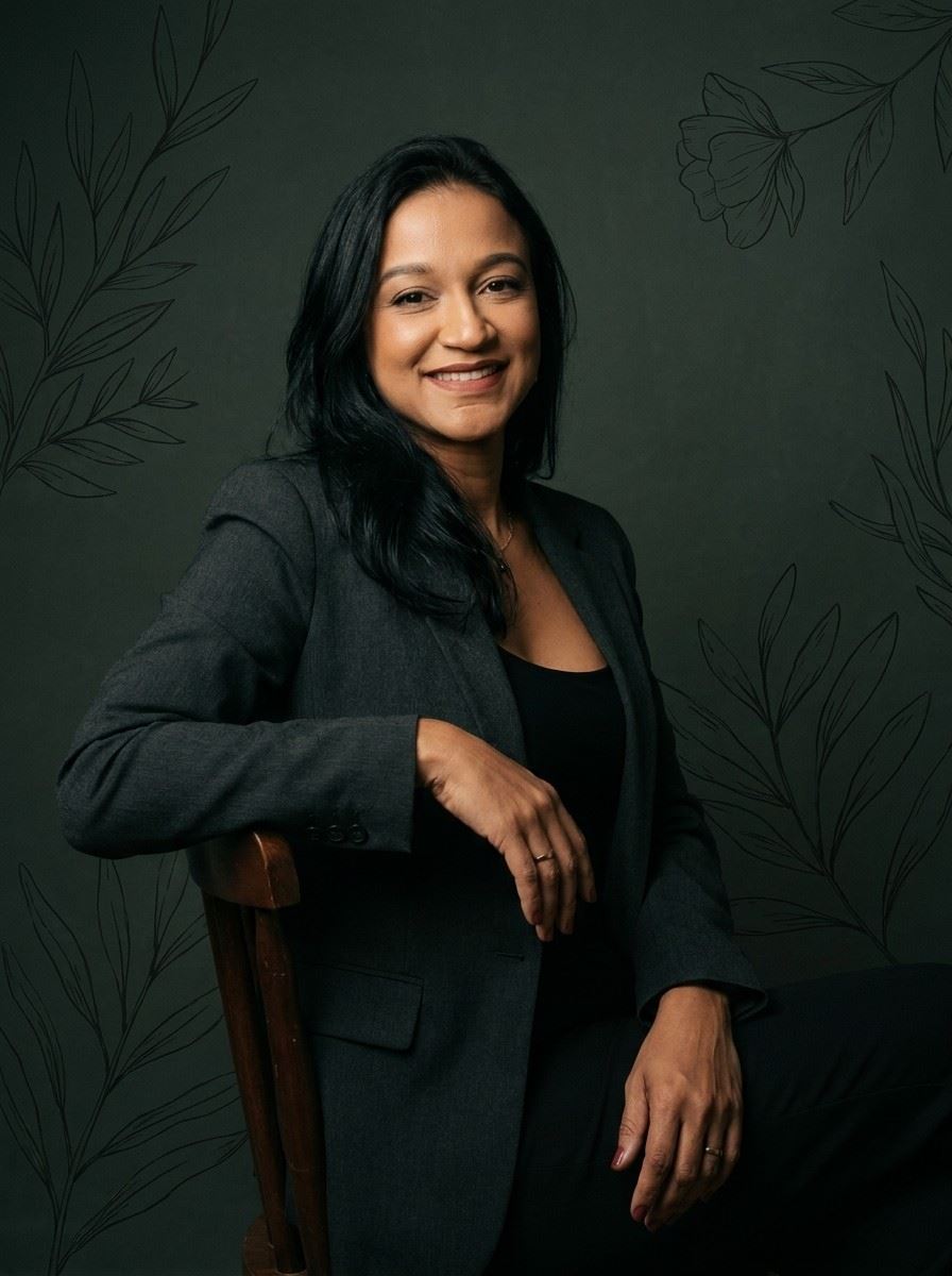 Retrato de Ana Oliveira, sorrindo, de blazer cinza, sentada diante de parede escura com desenhos de folhas