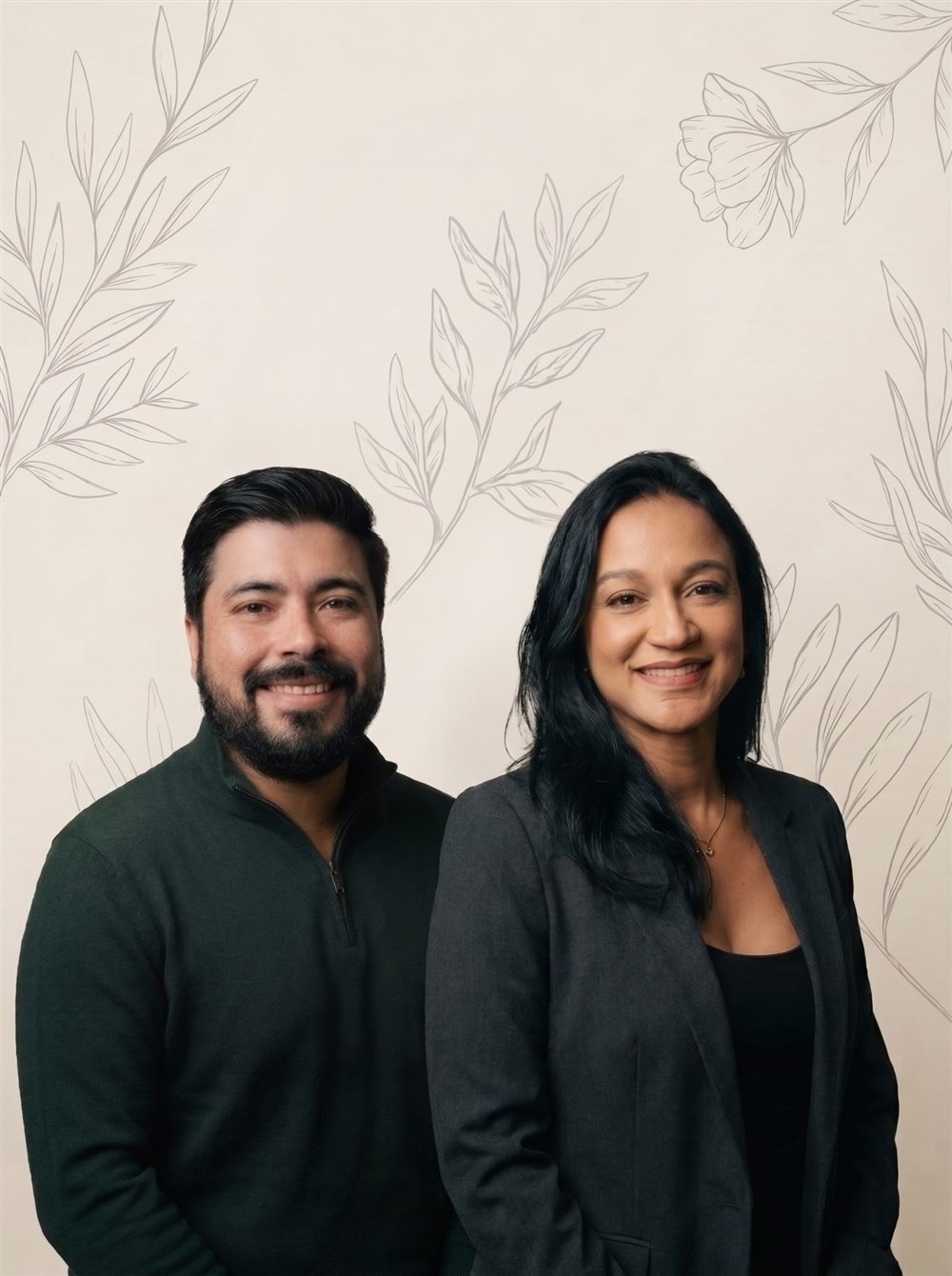 Ana Oliveira e Denis Faria, lado a lado, sorrindo, diante de uma parede creme com desenhos de folhas